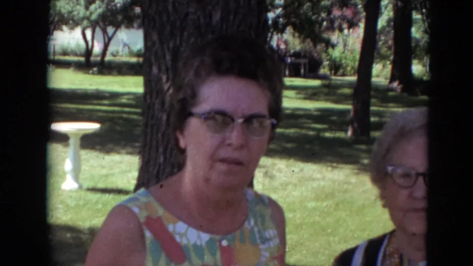 In a sun-dappled Lincoln, Nebraska outdoor setting, a woman in a colorful sleeveless dress faces the camera with another older woman partly in frame.