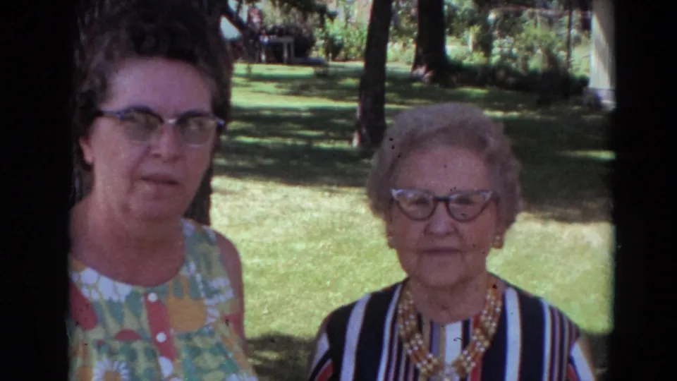 Two women in 1974 Lincoln, Nebraska home-movie footage pose outdoors, one in a colorful dress and one in a striped top with a beaded necklace.