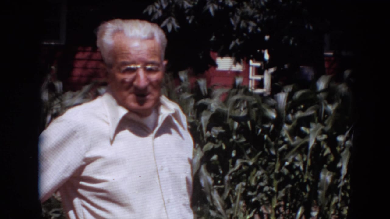 Older white-haired man in glasses standing before corn plants and a red building.