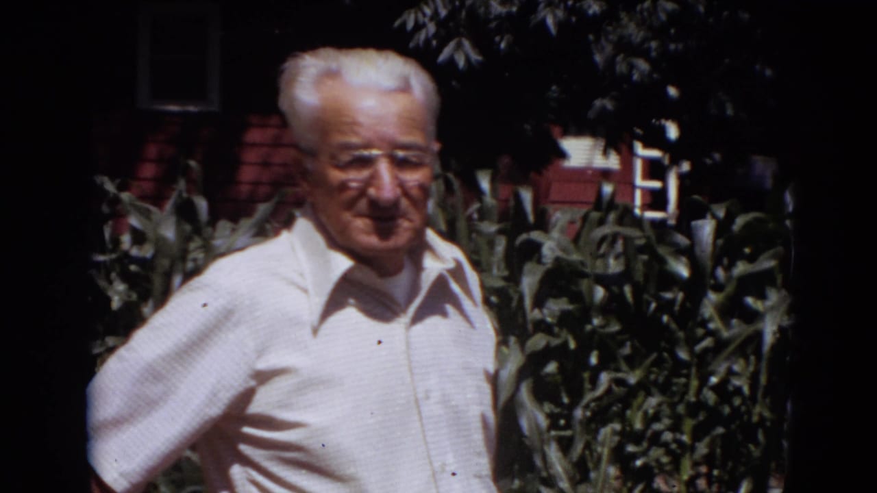 The man remains near the garden, framed from the waist up against corn leaves and a red-sided structure.