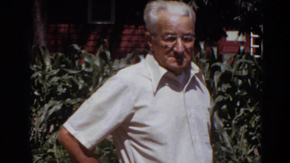 The man stands at the garden edge in Lincoln, Nebraska, with bright sun on his shirt and corn leaves behind him.