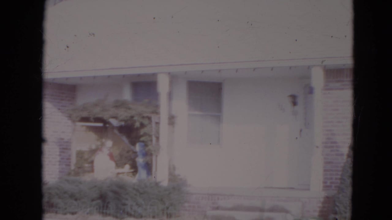 Brick-and-white house front with porch posts, shrubs, arbor, and two distant figures near the entry.