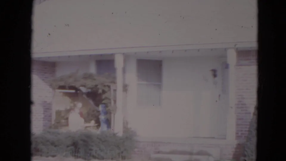 1974 Lincoln, Nebraska 8mm frame showing a suburban house front with porch posts, shrubs, arbor, and distant figures near the doorway.