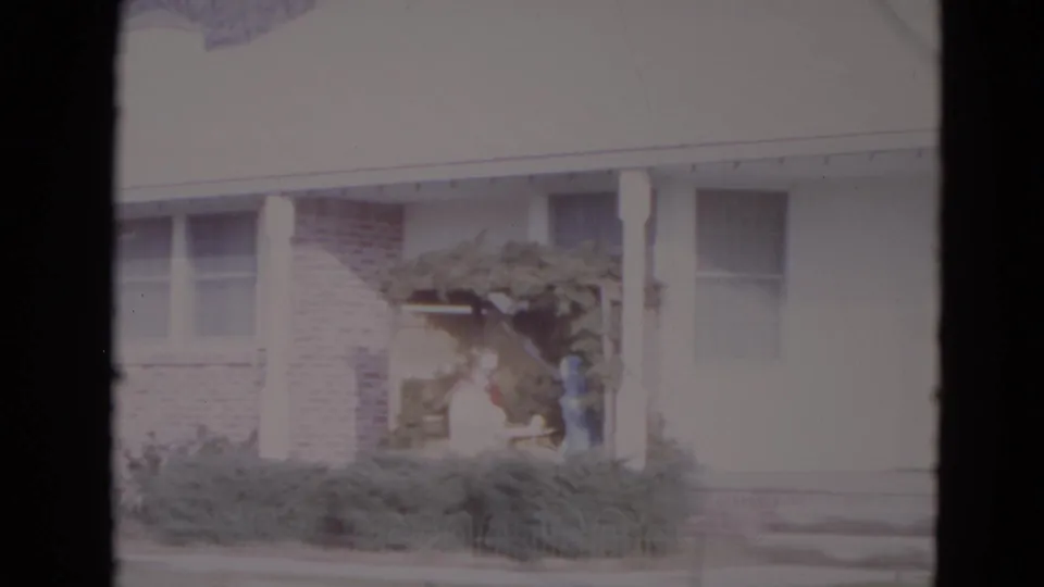 1974 Lincoln, Nebraska home-movie frame of the porch side of a suburban house with shrubs, arbor, and distant figures at the entry.