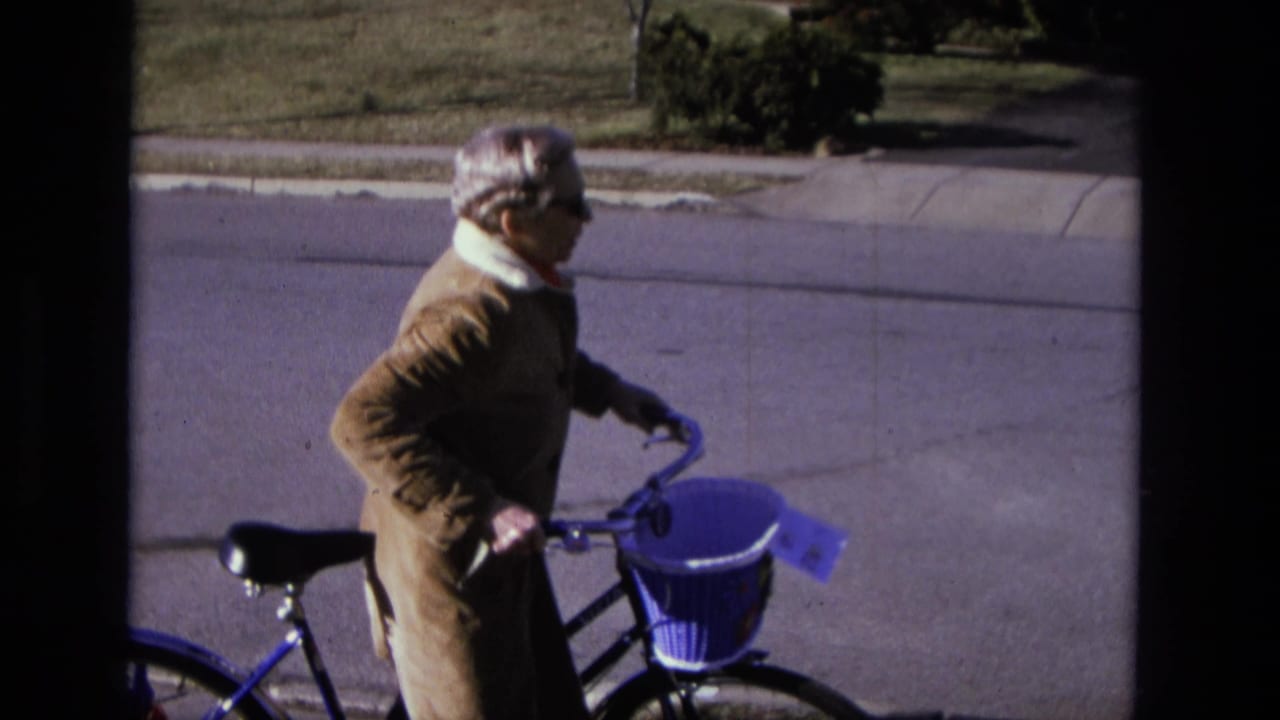 Elderly woman in a brown coat holds a bicycle with a purple front basket beside a residential street.