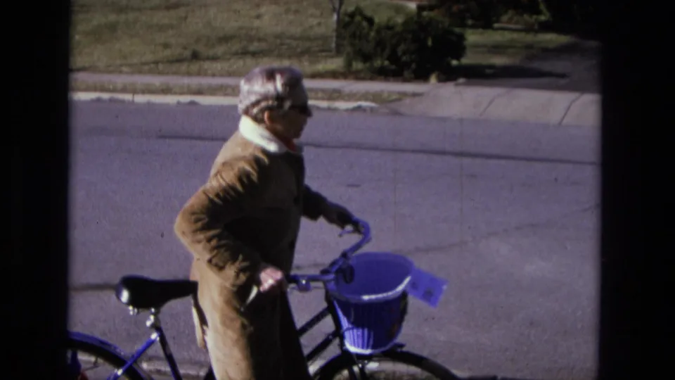 In 1975 California home-movie footage, an elderly woman in a brown coat stands with a blue bicycle and purple basket near a sidewalk.