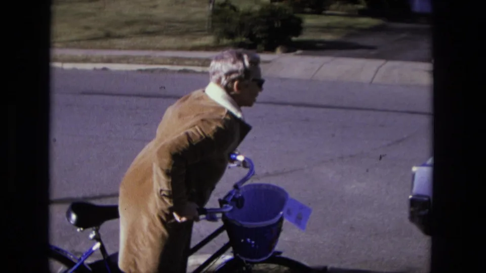 The elderly woman bends toward the bicycle handlebars on a California sidewalk, with a vehicle partly visible at frame right.