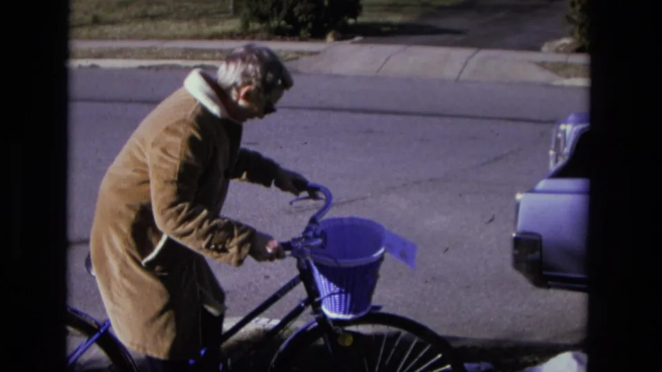 The home-movie frame shows the woman adjusting or steadying her bicycle beside the street, with a car cropped at the right edge.