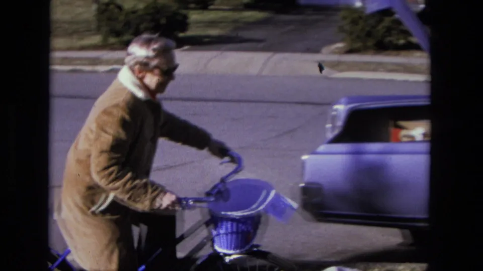 An elderly woman holds her bicycle in a 1975 California suburban scene while a station wagon moves through the background.