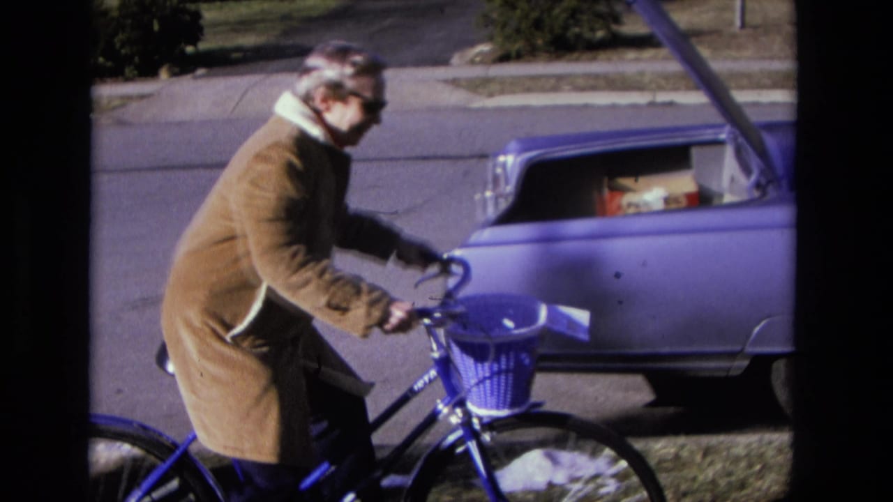 Adult in a brown coat rides a blue bicycle beside a parked blue sedan with its trunk open.