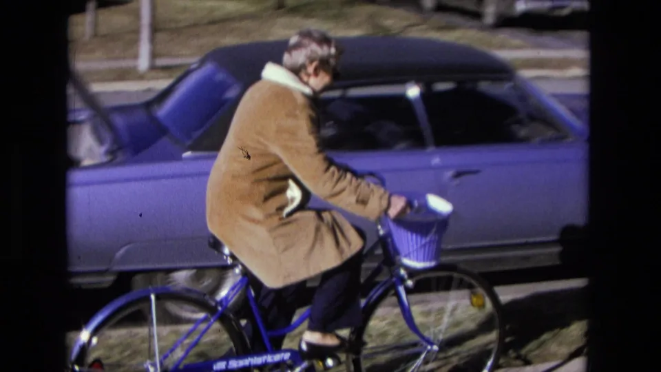 Adult rider in a long brown coat passes a parked blue sedan on a blue bicycle with a purple front basket in 1975 California footage.