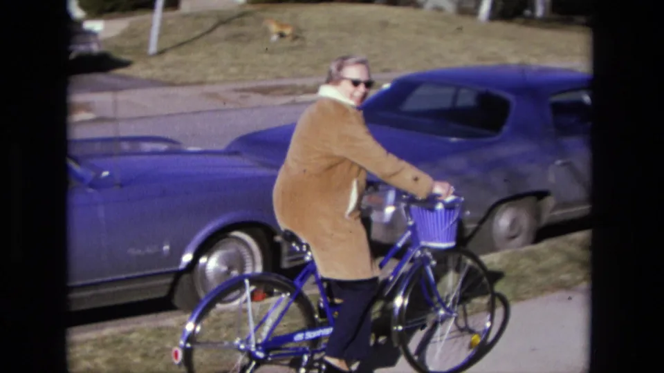 The cyclist briefly turns toward the camera while riding a blue basket bicycle past a vintage blue sedan in suburban California.