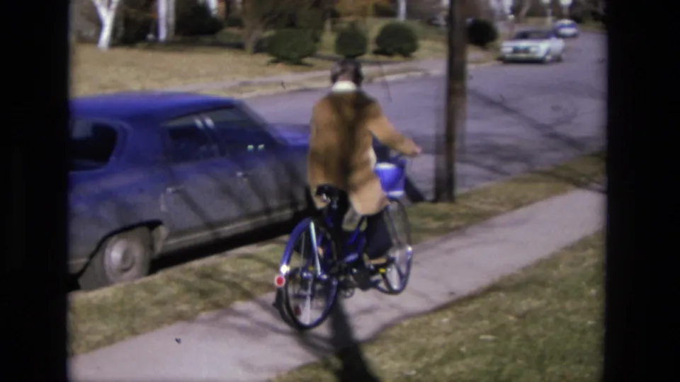 The rider moves away down a California suburban sidewalk on a blue bicycle, with lawns, parked cars, and a quiet street visible.