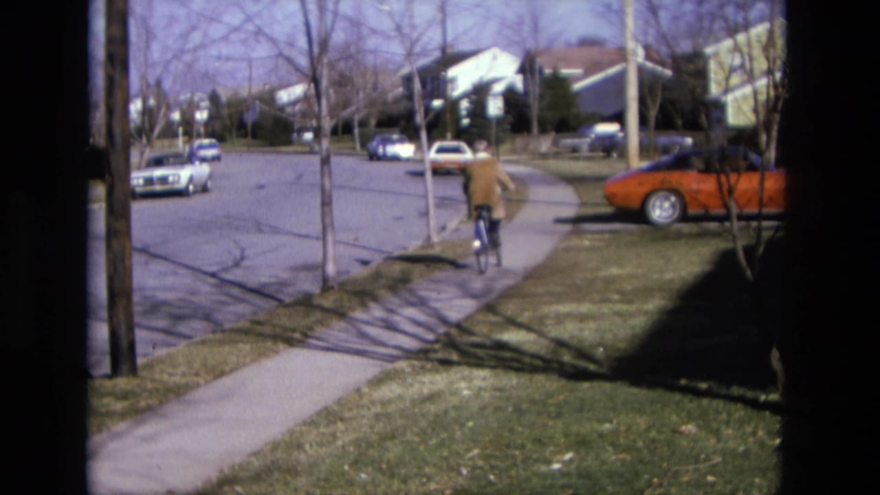 Person riding a bicycle along a suburban sidewalk past parked cars and houses.
