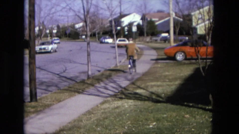 In 1975 California home-movie footage, a bicyclist rides away along a sidewalk beside lawns, bare trees, and parked cars.
