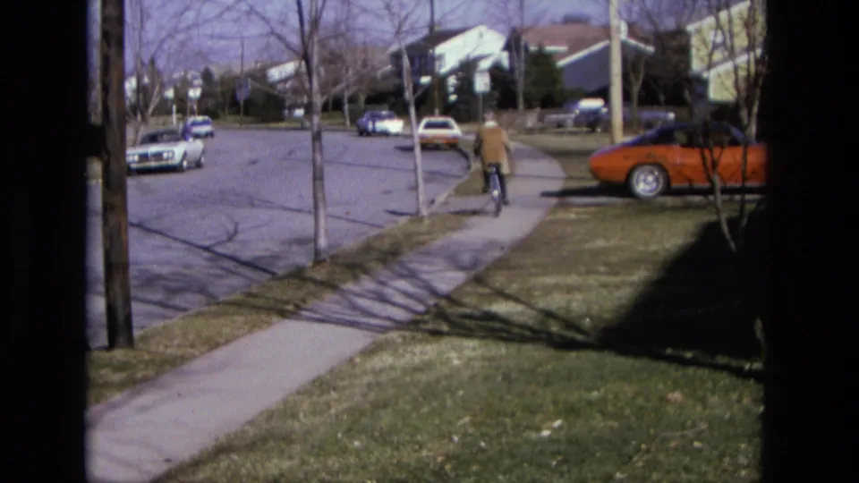 The bicyclist rides farther down a California suburban sidewalk, with an orange coupe, sedans, lawns, and houses nearby.