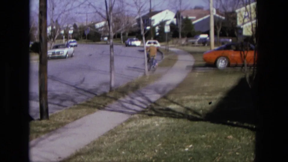 A distant rider in a brown coat pedals along the sidewalk toward a residential street with cars, trees, and houses visible.