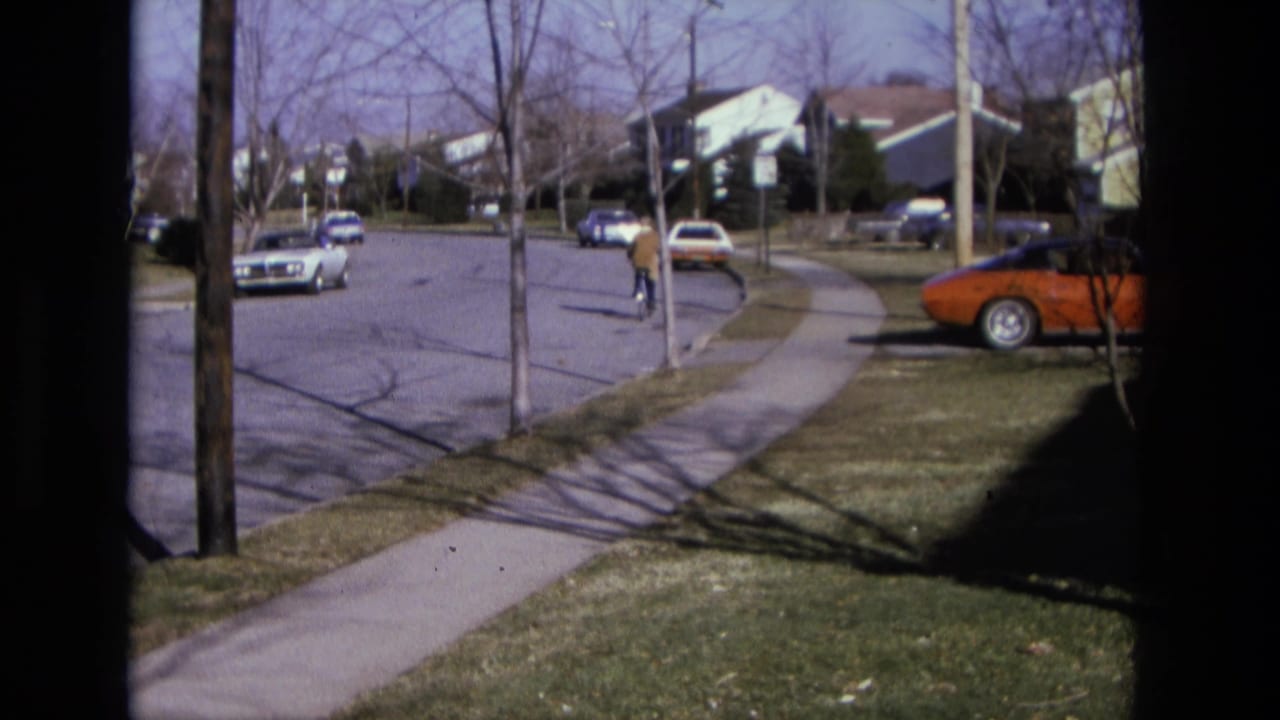 1975 California suburban street seen from a fixed window-like viewpoint, with parked cars and a distant cyclist on the road.