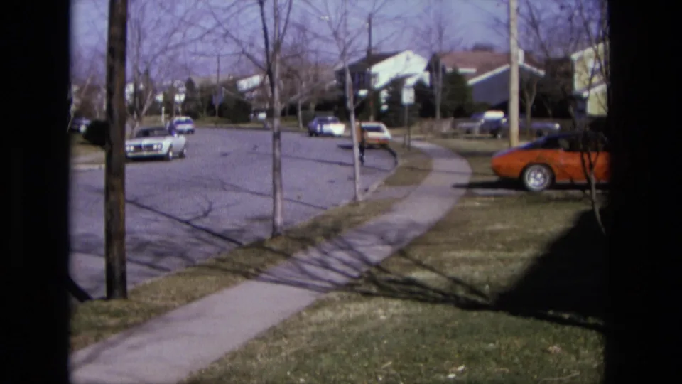 Suburban California street in 1975 with a distant cyclist, curbside cars, sidewalks, lawns, and bare trees under bright daylight.