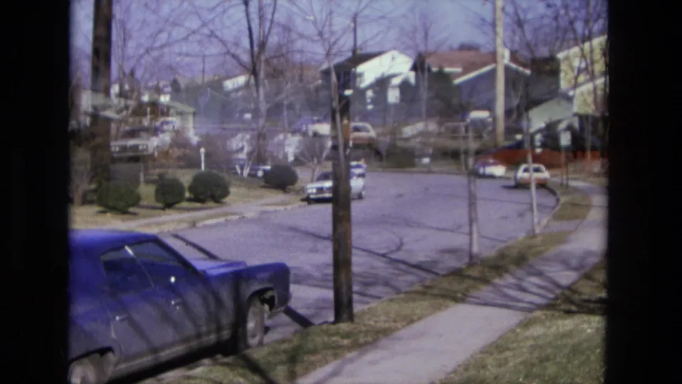 1975 California neighborhood view with a blue car in the foreground, parked vehicles, houses, lawns, and bare trees on a sunny street.