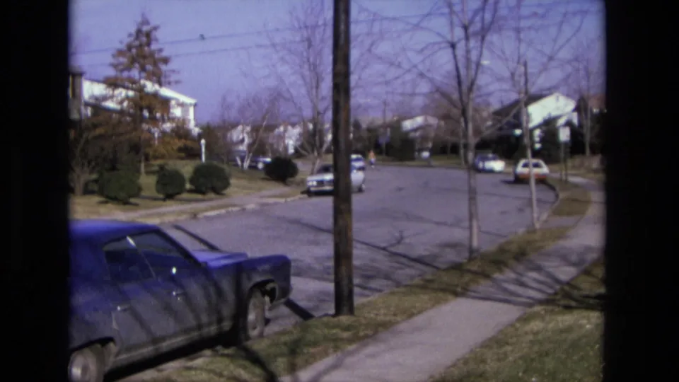 Suburban California street scene from 1975 showing a blue car in the foreground, distant parked cars, sidewalks, and bare winter trees.