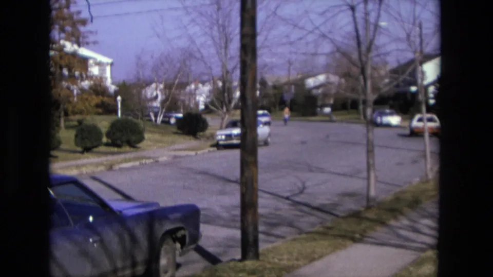 1975 California home-movie frame with a bicyclist moving down a suburban road between parked cars, sidewalks, lawns, and houses.