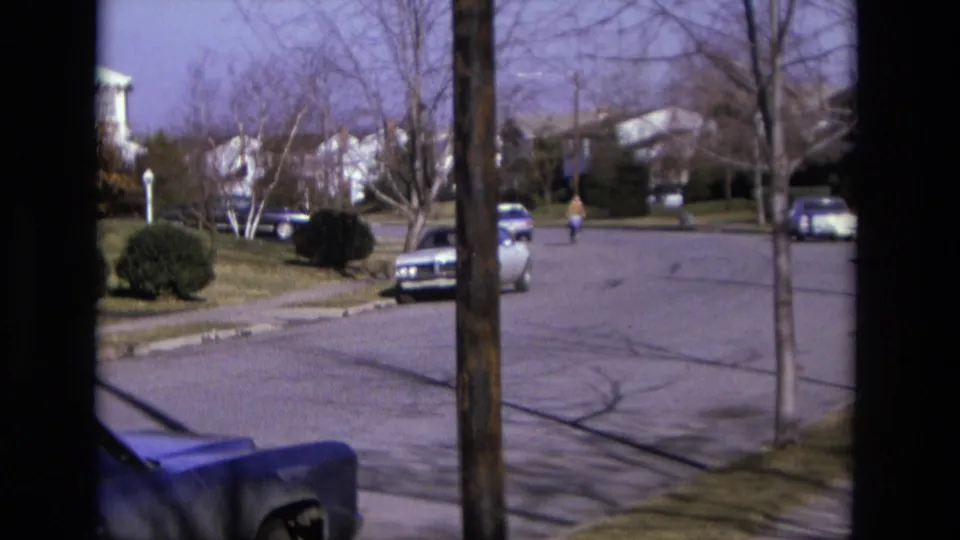 1975 California 8mm still showing a bicyclist nearer in the roadway, with parked cars, bare trees, sidewalks, and suburban houses.