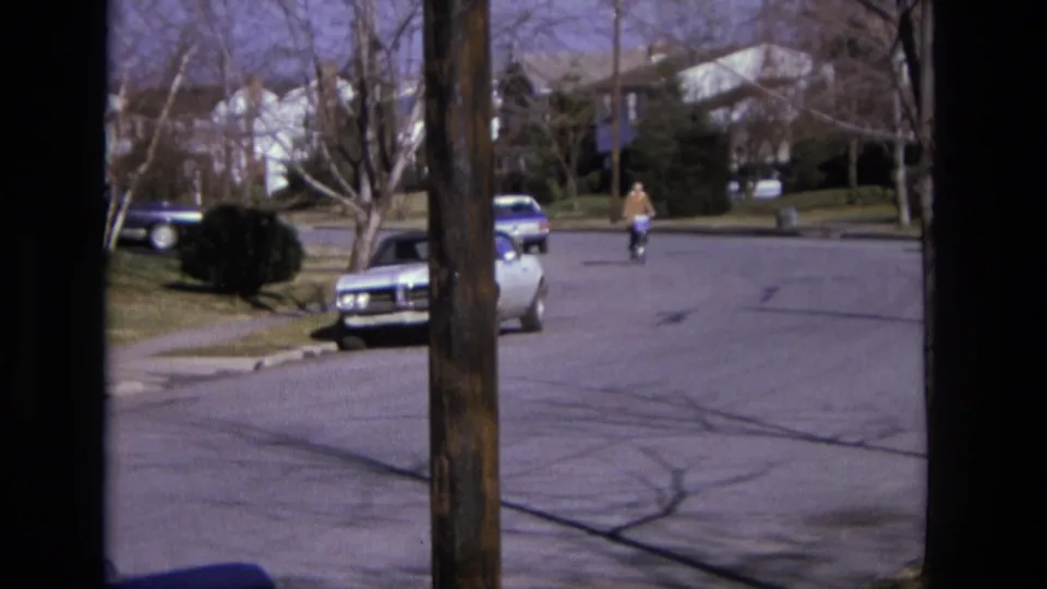 1975 California frame of a person riding a bicycle along a quiet suburban street, partly framed by a foreground utility pole.