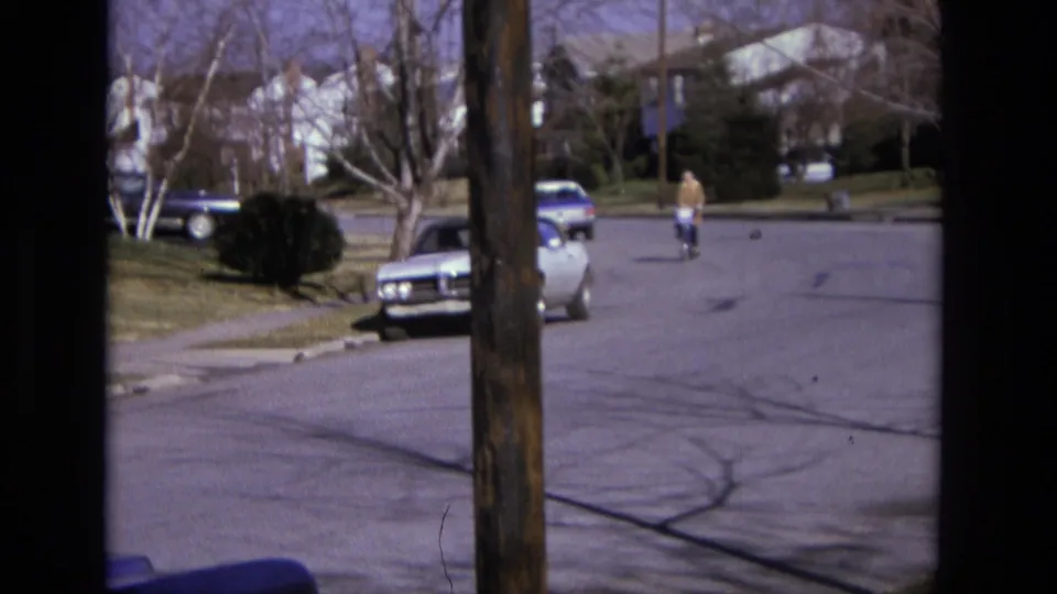 1975 California home-movie frame with a distant cyclist, curbside cars, residential houses, bare trees, and visible street cracks.