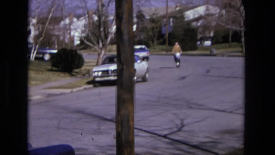 1975 California 8mm frame showing a cyclist nearer the camera on a quiet suburb street, with cars, houses, trees, and a pole visible.