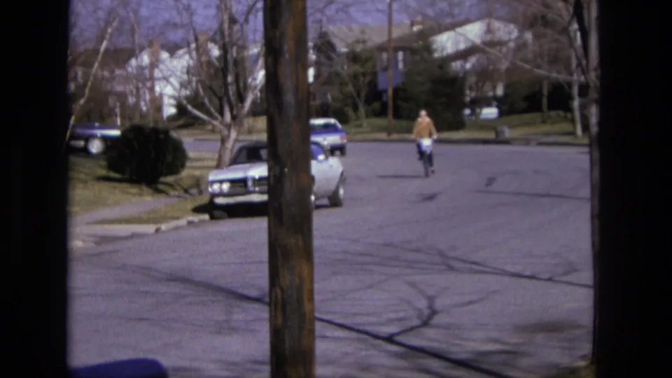 1975 California home-movie frame of a cyclist on a suburban street with parked cars, bare trees, houses, and dark window framing.