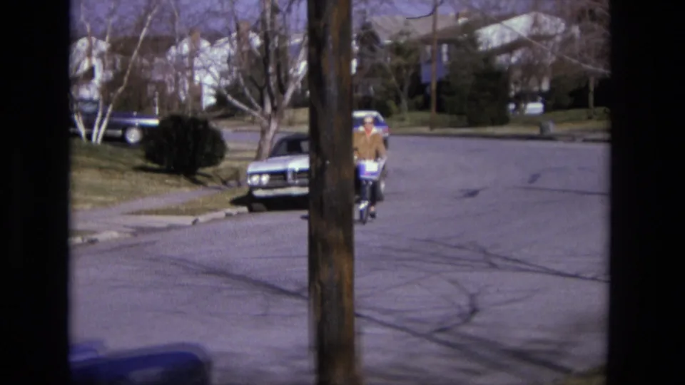 The cyclist moves closer through the residential block, with 1970s-era cars, lawns, and bare trees visible in the background.