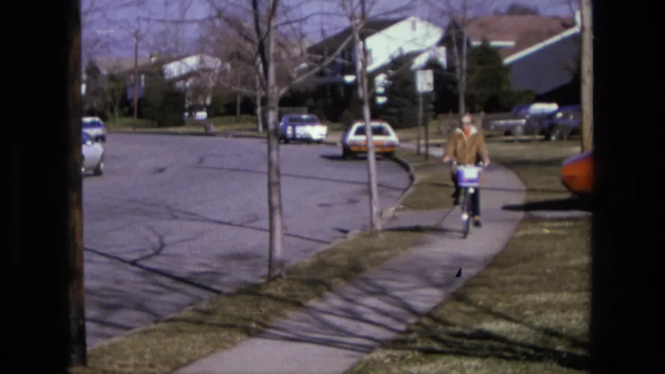A cyclist in casual clothing rides along the sidewalk in a California suburb, passing lawns, parked cars, and leafless street trees.