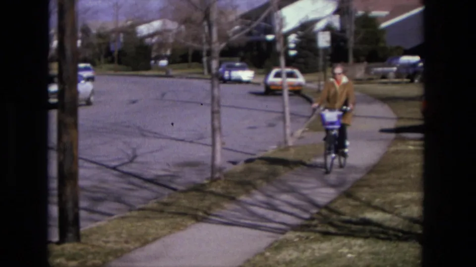 The cyclist continues down the sidewalk past lawns and parked cars, with residential houses and dark window framing still visible.