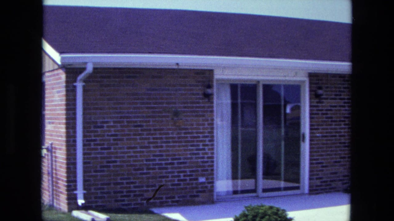 Red brick house exterior with sliding glass doors, patio, roofline, lawn, and shrub.