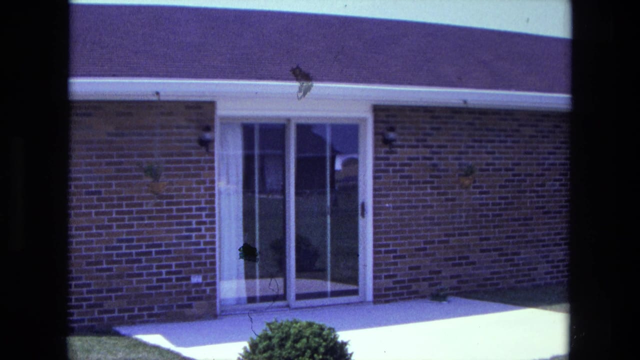 1975 California home-movie frame of a brick house facade with glass doors, white trim, patio slab, roofline, and lawn.