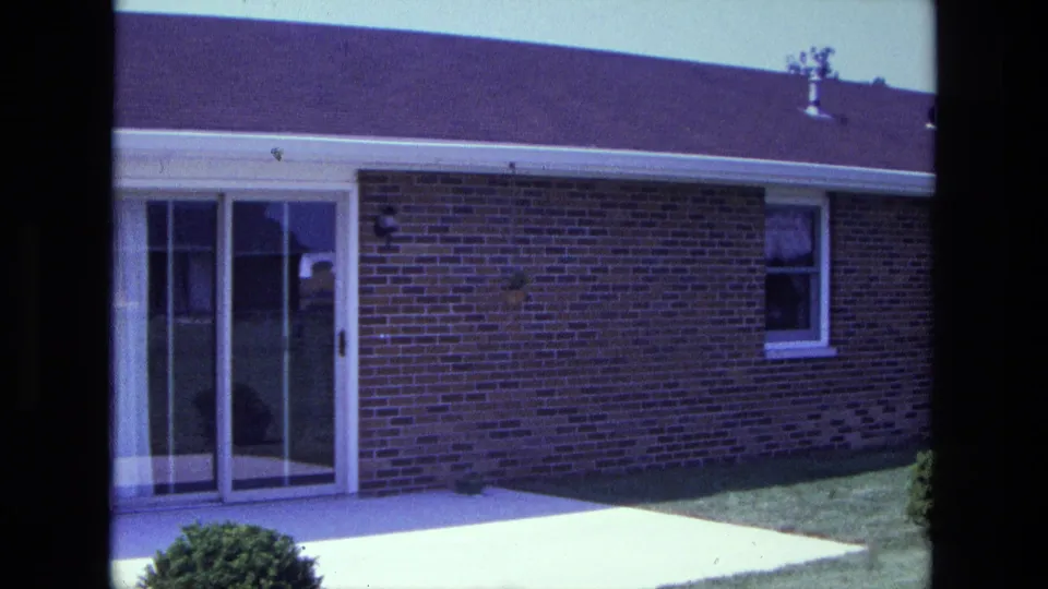 1975 California frame showing the brick house as the pan continues past sliding glass doors toward a small window and lawn.