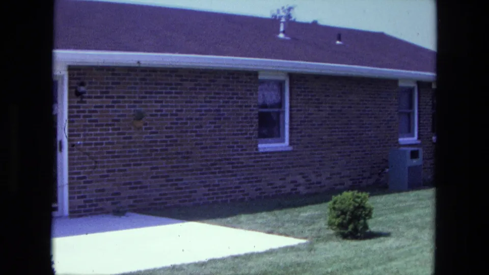 1975 California home-movie frame of a red brick residential wall with small windows, trimmed lawn, shrub, patio, and utility box.