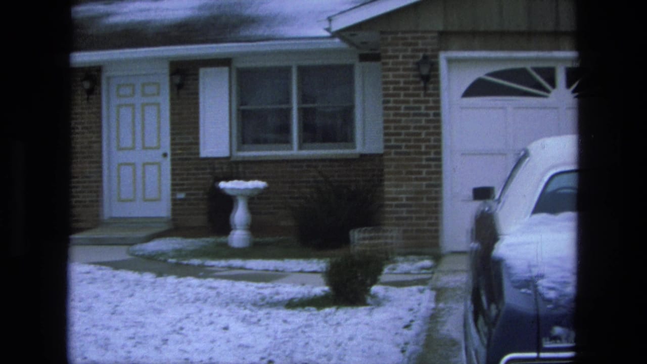 1975 California home-movie frame showing a brick ranch-style house, snowy lawn, white birdbath, and part of a blue sedan in the driveway.