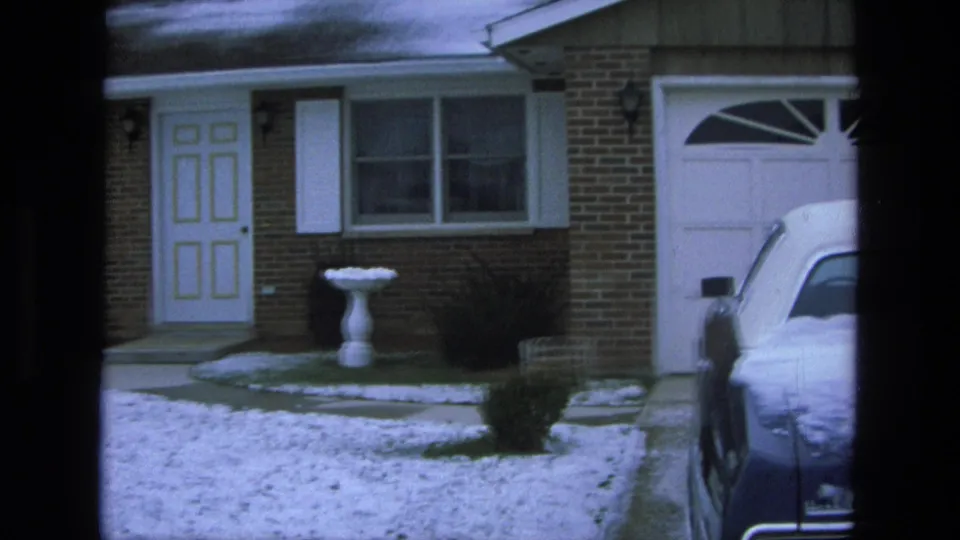 1975 California home-movie frame showing a brick ranch-style house, snowy lawn, white birdbath, and part of a blue sedan in the driveway.