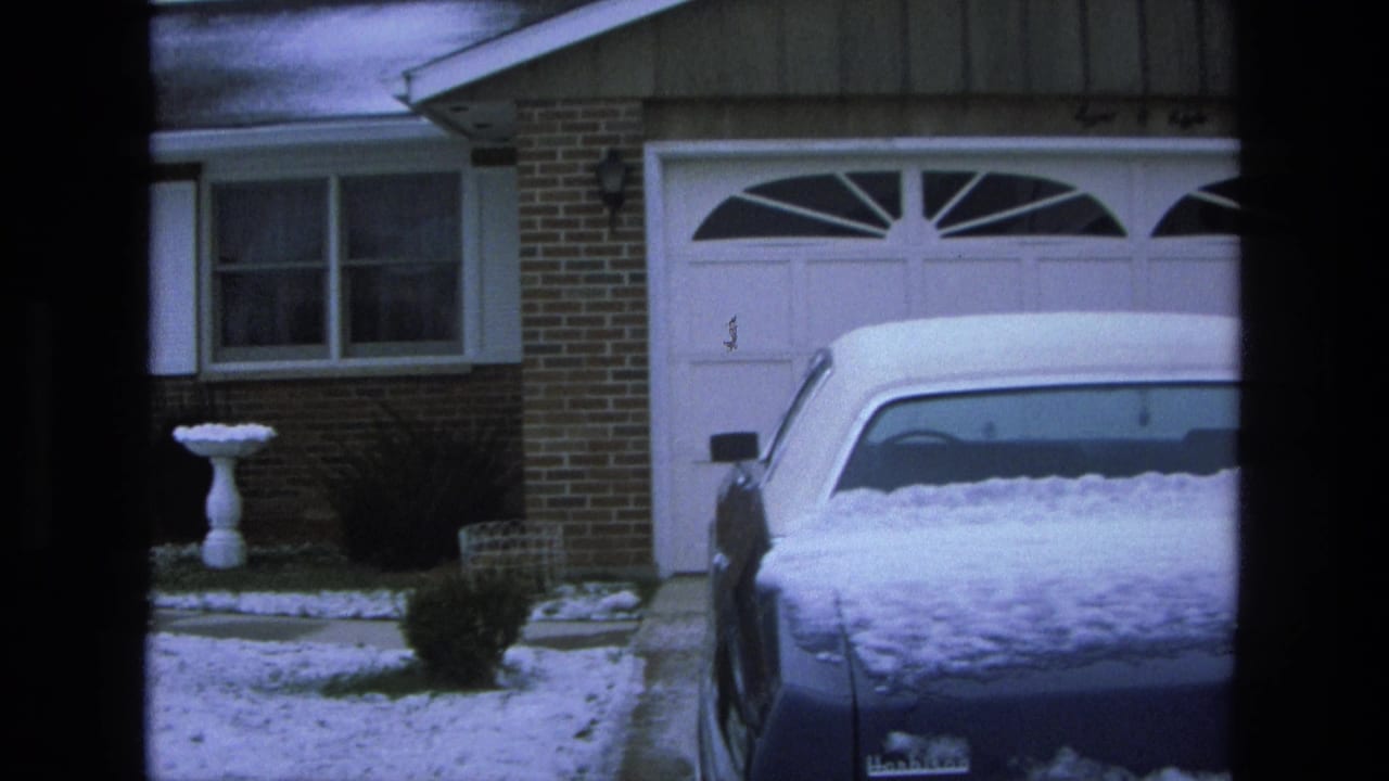 1975 California suburban winter view with snow on the driveway, a white garage door, birdbath, shrubs, and a parked blue sedan.