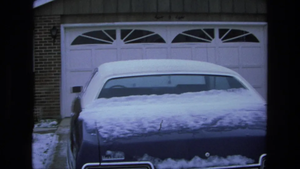 Close 1975 home-movie view of a snow-covered blue sedan backed into a suburban driveway before white garage doors.