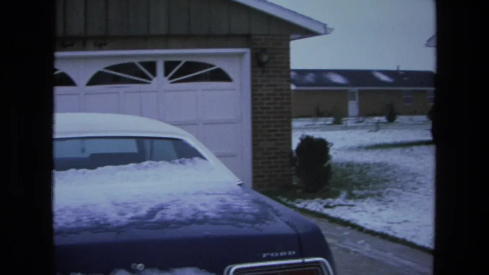 1975 California frame showing a snow-covered blue Ford LTD beside a white garage, with snowy suburban lawns and houses in the background.