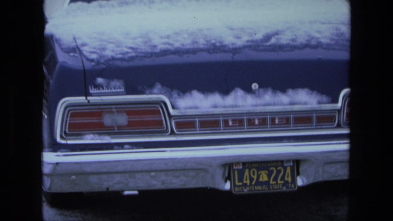 Rear of a blue Ford LTD sedan with snow on the trunk and roof, parked before a garage.
