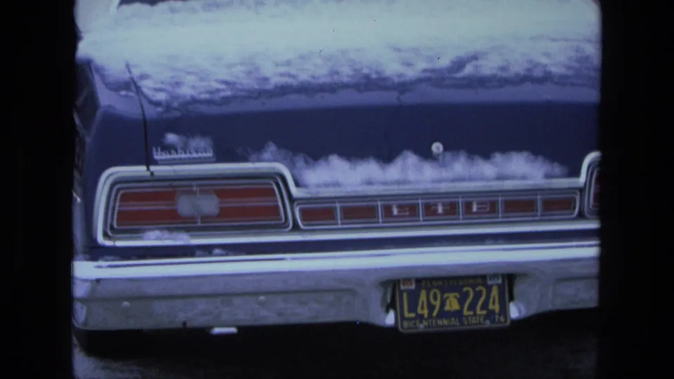 1975 California 8mm frame showing the rear of a blue Ford LTD sedan with snow across the trunk, roof, chrome bumper, and black plate.