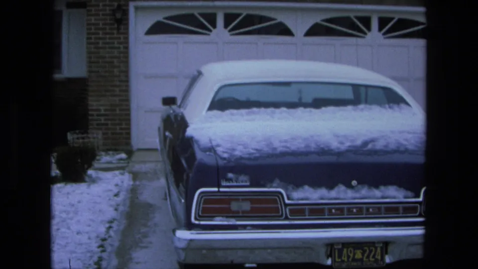 1975 California 8mm frame showing a blue Ford LTD with snow on it, parked outside a brick residential garage.