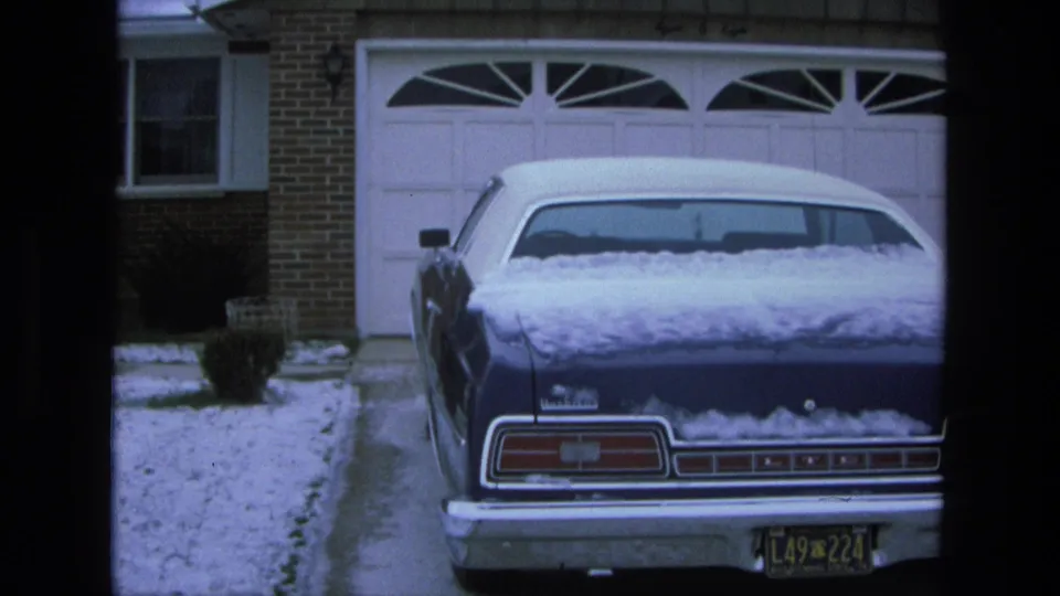 1975 California frame showing the rear of a blue Ford LTD with snow on the roof and trunk in a suburban driveway.