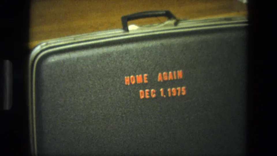 1975 California 8mm still with the suitcase filling most of the frame and orange Home Again date text visible.