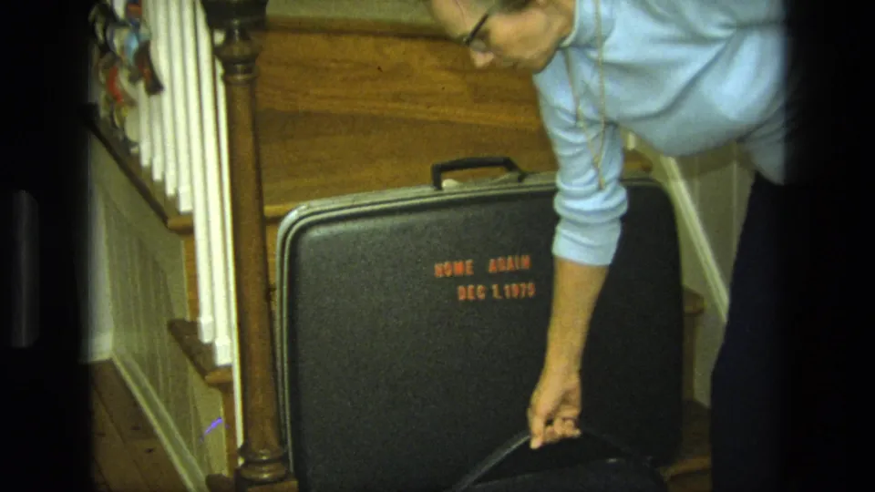A person in a light blue sweater reaches toward the black suitcase on a 1975 California home staircase.