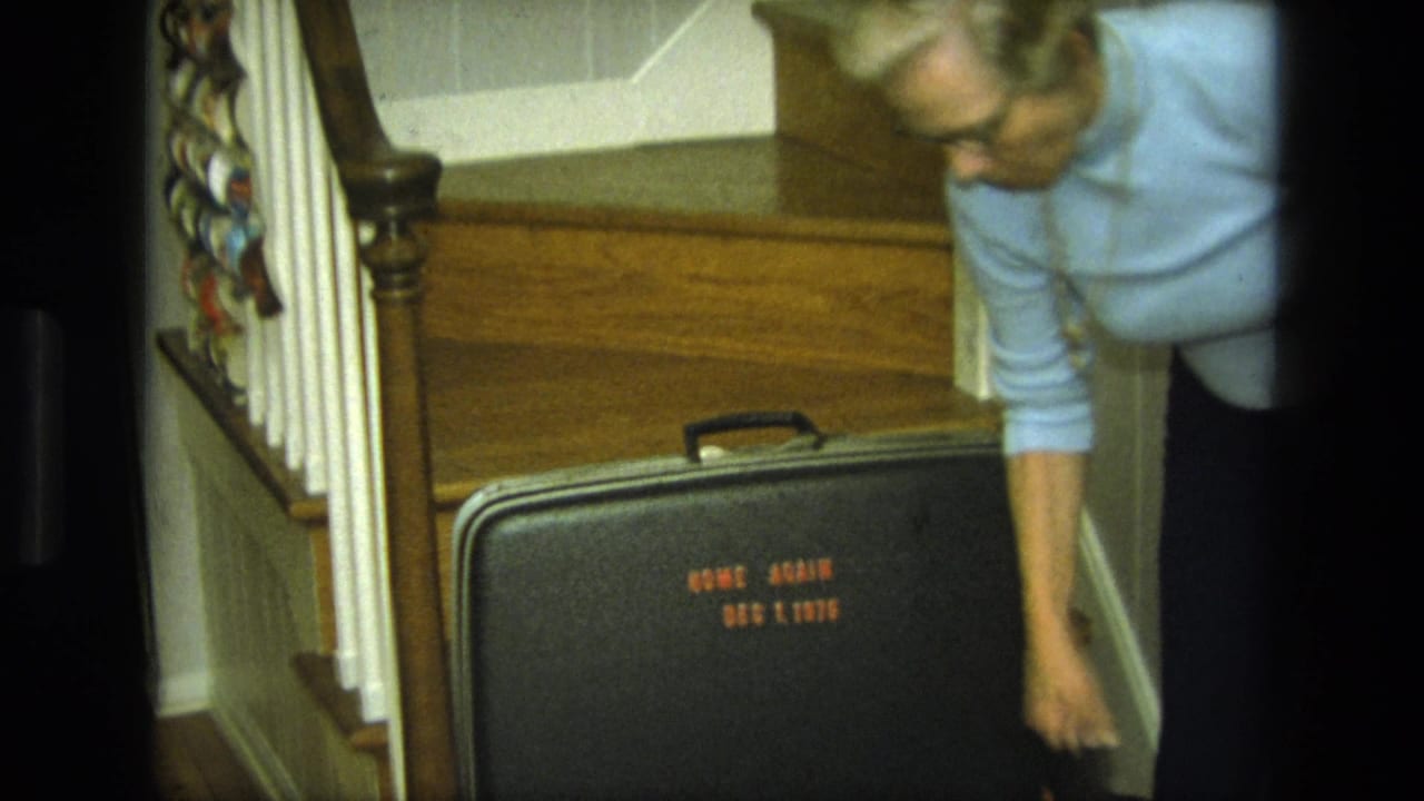 Blond woman bends beside a black suitcase at the bottom of a wooden staircase.